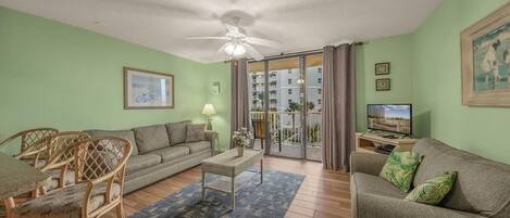 A living room with a sofa, a coffee table, and a dining table with chairs. There is a ceiling fan, a television, and a window with curtains.