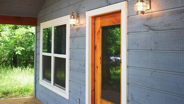 Una terraza de madera que conduce a una casa con un ventanal y una puerta, y dos luces fijadas a la pared.