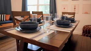 Una mesa de comedor dispuesta con cuencos y vasos oscuros, rodeada de sillas, en una habitación con ventanal y decoración en la pared.