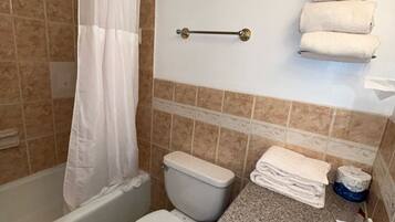 Un baño con inodoro, lavabo y bañera. Hay toallas apiladas en la pared y un rollo de papel higiénico en la encimera.
