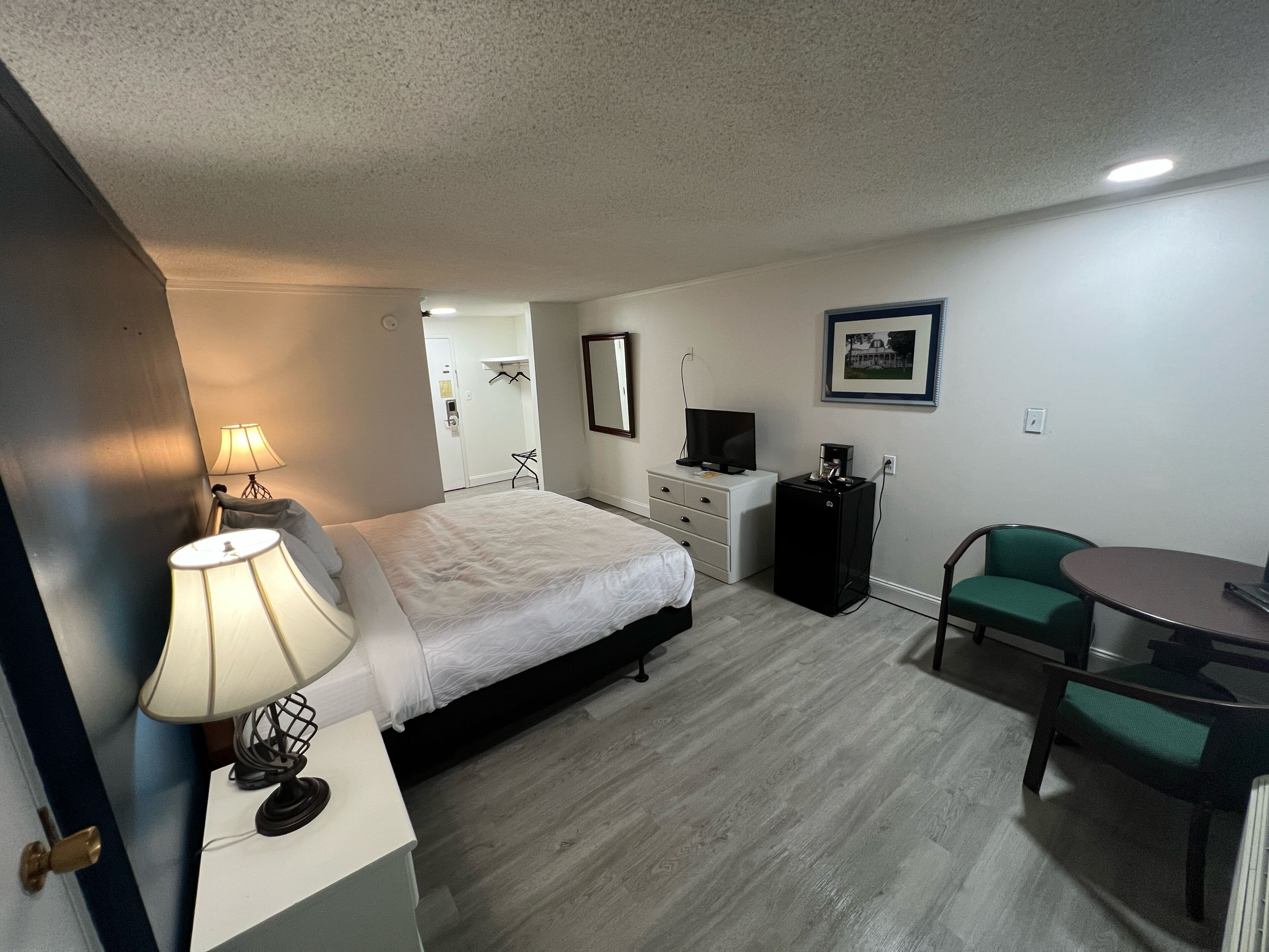 A hotel room with a bed, bedside lamp, desk with a lamp, a television, a coffee machine, a chair, and a small table.