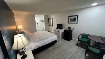 A hotel room with a bed, bedside lamp, desk with a lamp, a television, a coffee machine, a chair, and a small table.