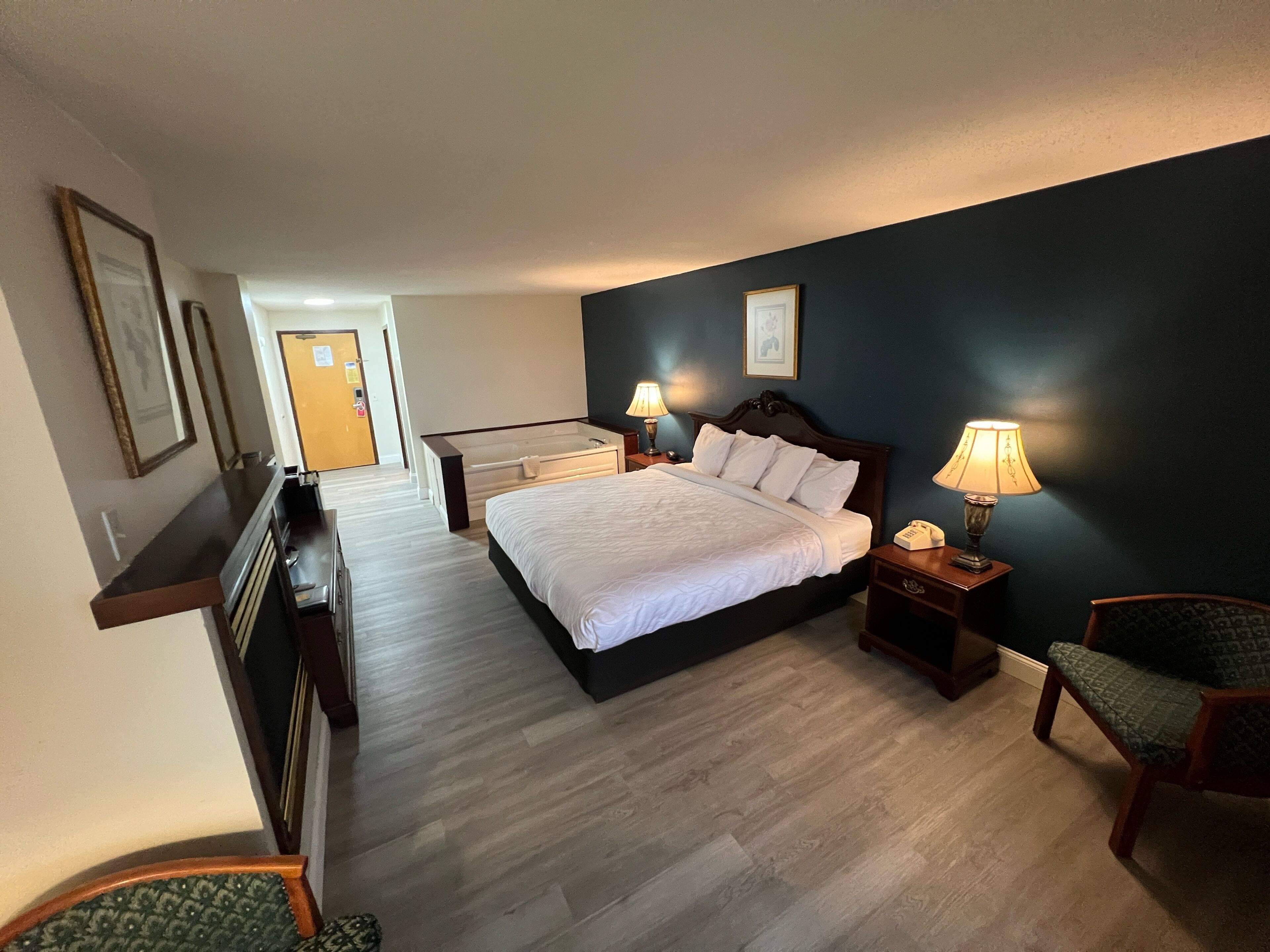 A hotel room with a large bed, bedside lamps, a chair, a desk, and a view of the hallway.