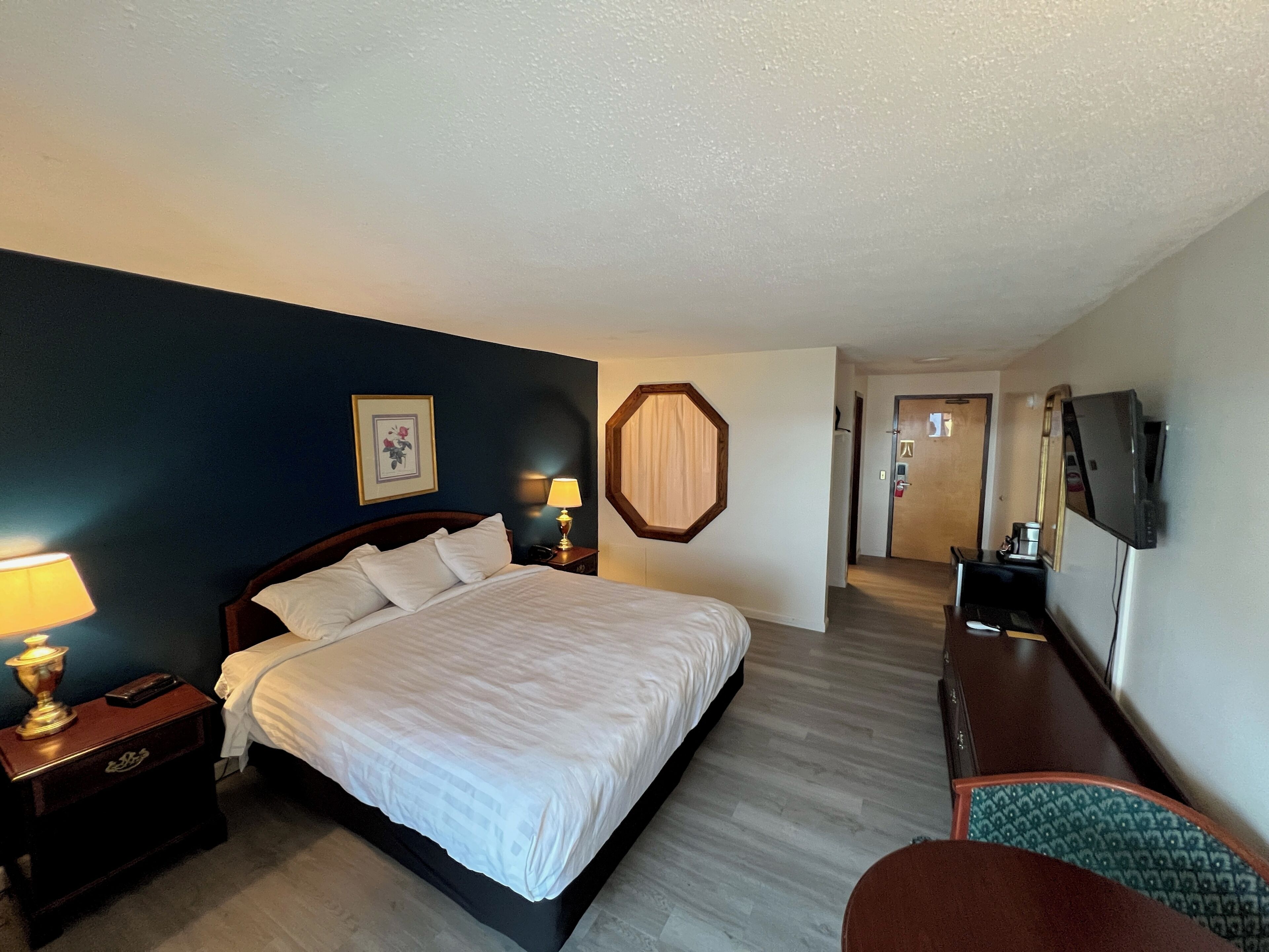 A hotel room with a bed, bedside lamps, a wooden nightstand, a framed picture on the wall, a mirror, a television mounted on the wall, and a door leading to another room.