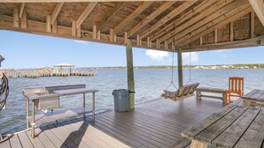 A wooden deck with a bench, a table, and a hanging chair overlooking a body of water. - Lagoon Run 105 (Gulf Shores)