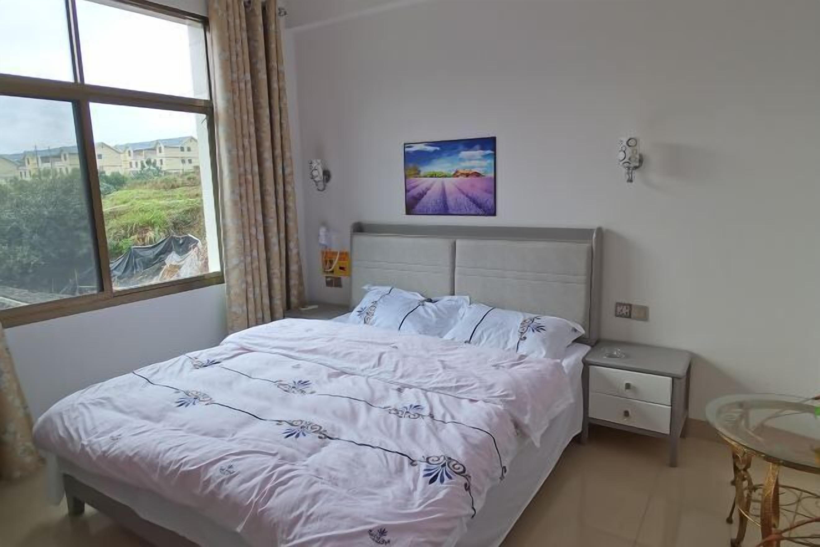 Un dormitorio con cama, mesita de noche, mesa y ventana con vista a edificios y áreas verdes.