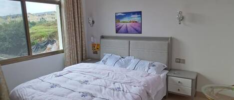 Un dormitorio con cama, mesita de noche, mesa y ventana con vista a edificios y áreas verdes.