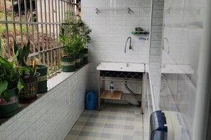 A small, tiled bathroom with a sink, a faucet, and a shelf above it. There are potted plants on the counter and a blue trash bin.