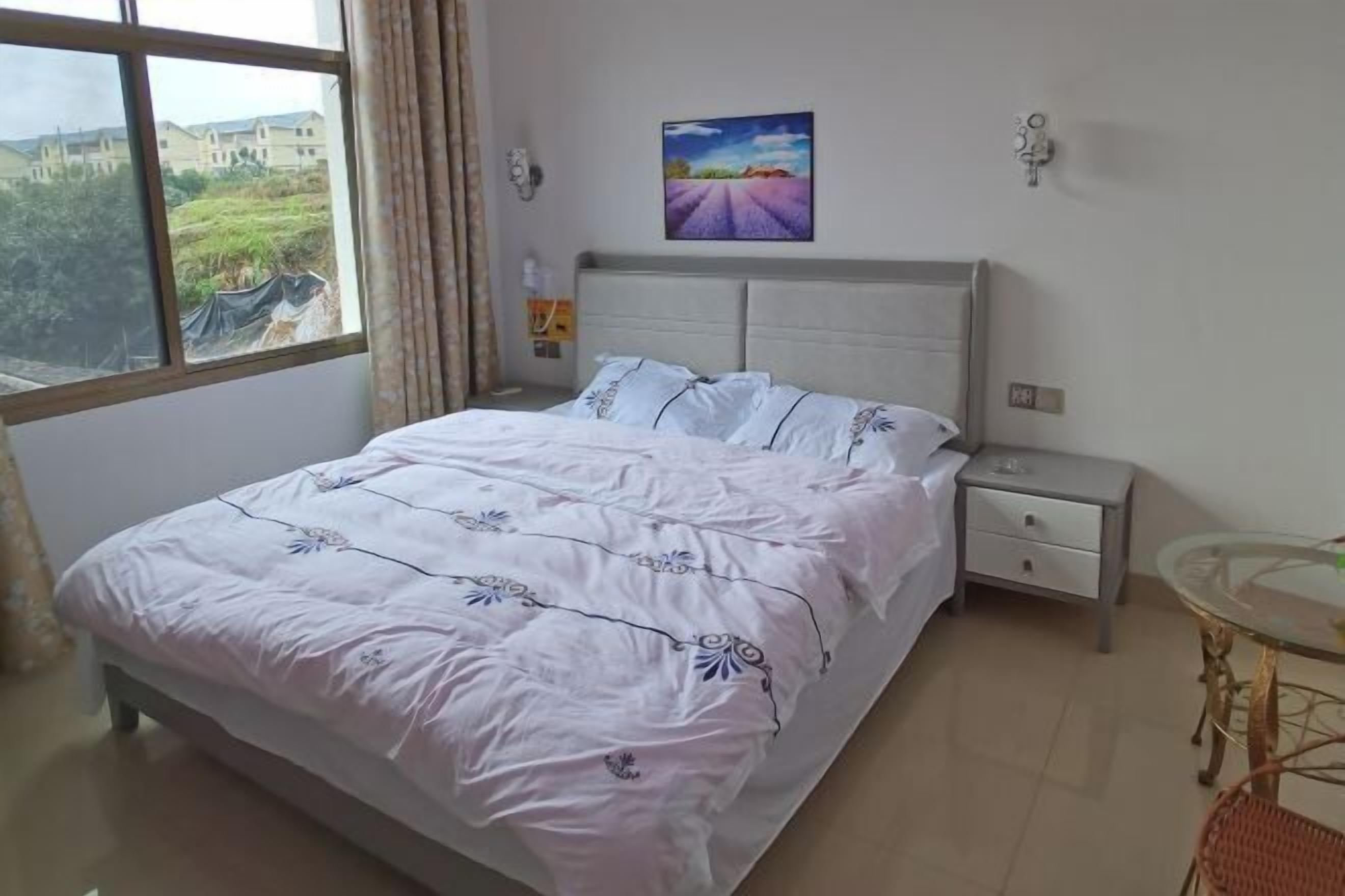 Une chambre à coucher avec un grand lit, une table de chevet, une petite table et une fenêtre donnant sur des bâtiments et de la verdure.