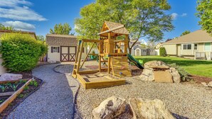 Un parque infantil con una estructura de madera para escalar y un tobogán, rodeado de un sendero de grava y follaje.