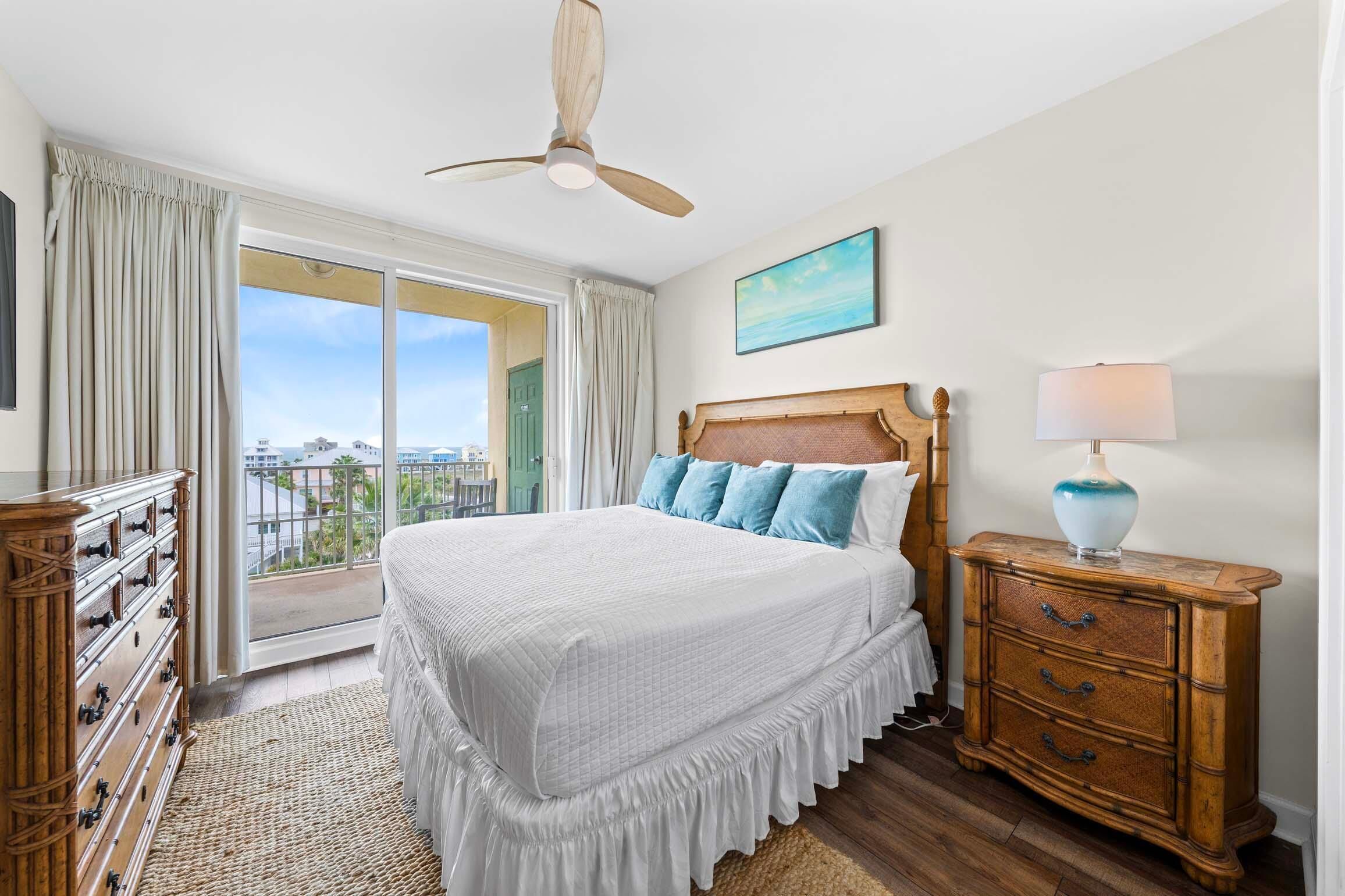 A bedroom with a bed, a dresser, a nightstand, a ceiling fan, a window with curtains, and a painting on the wall.