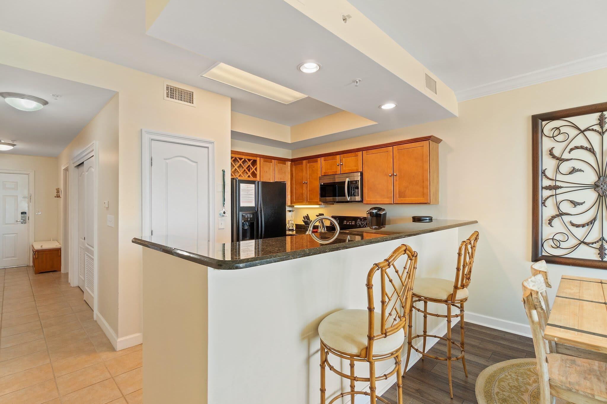 A kitchen with wooden cabinets, a stainless steel refrigerator, a microwave, and a bar area with two wicker chairs.