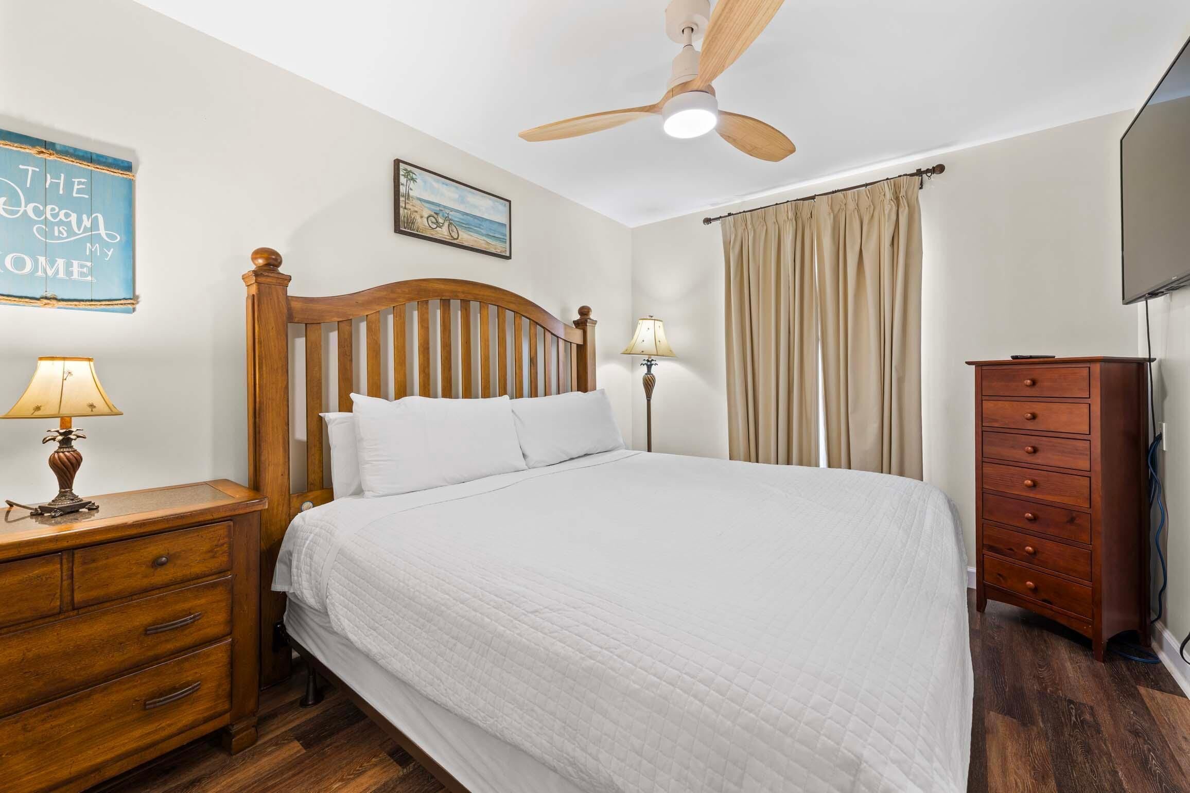 Un dormitorio con cama de madera, mesita de noche con lámpara, cómoda, ventilador de techo, adorno de temática playera y una pintura de playa.