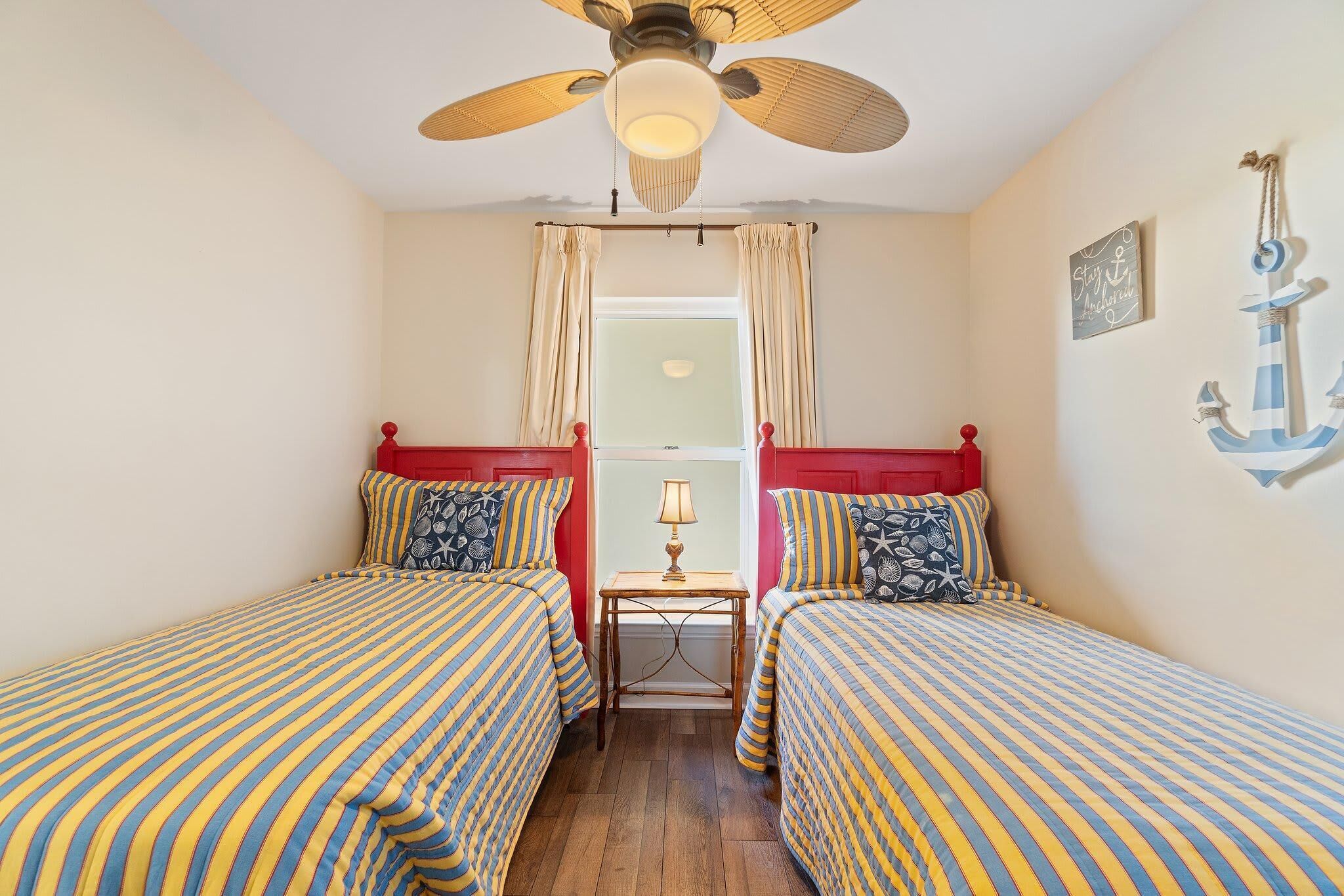 A room with two beds, a ceiling fan, a window with curtains, a small table with a lamp, and nautical-themed wall decor.