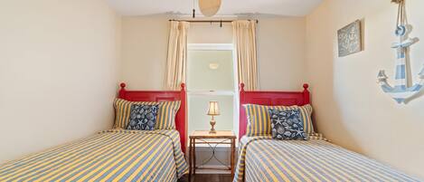 A room with two beds, a ceiling fan, a window with curtains, a small table with a lamp, and nautical-themed wall decor.