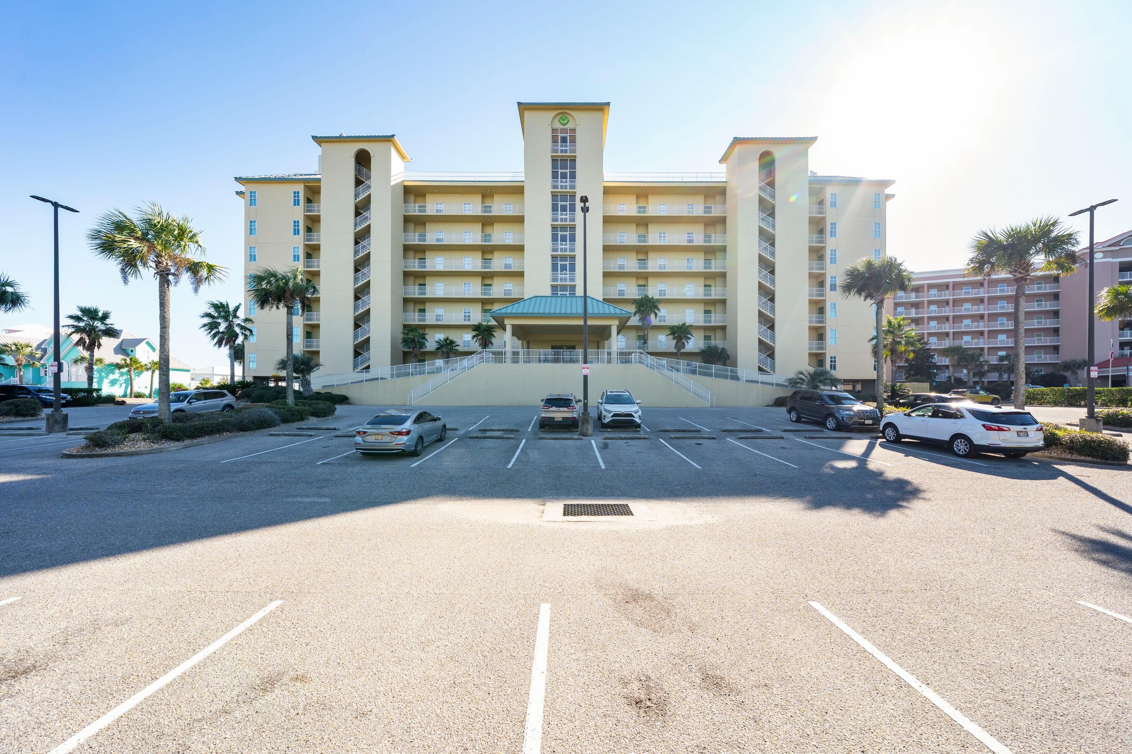 A multi-story hotel with a central entrance, parking lot, and surrounding palm trees.