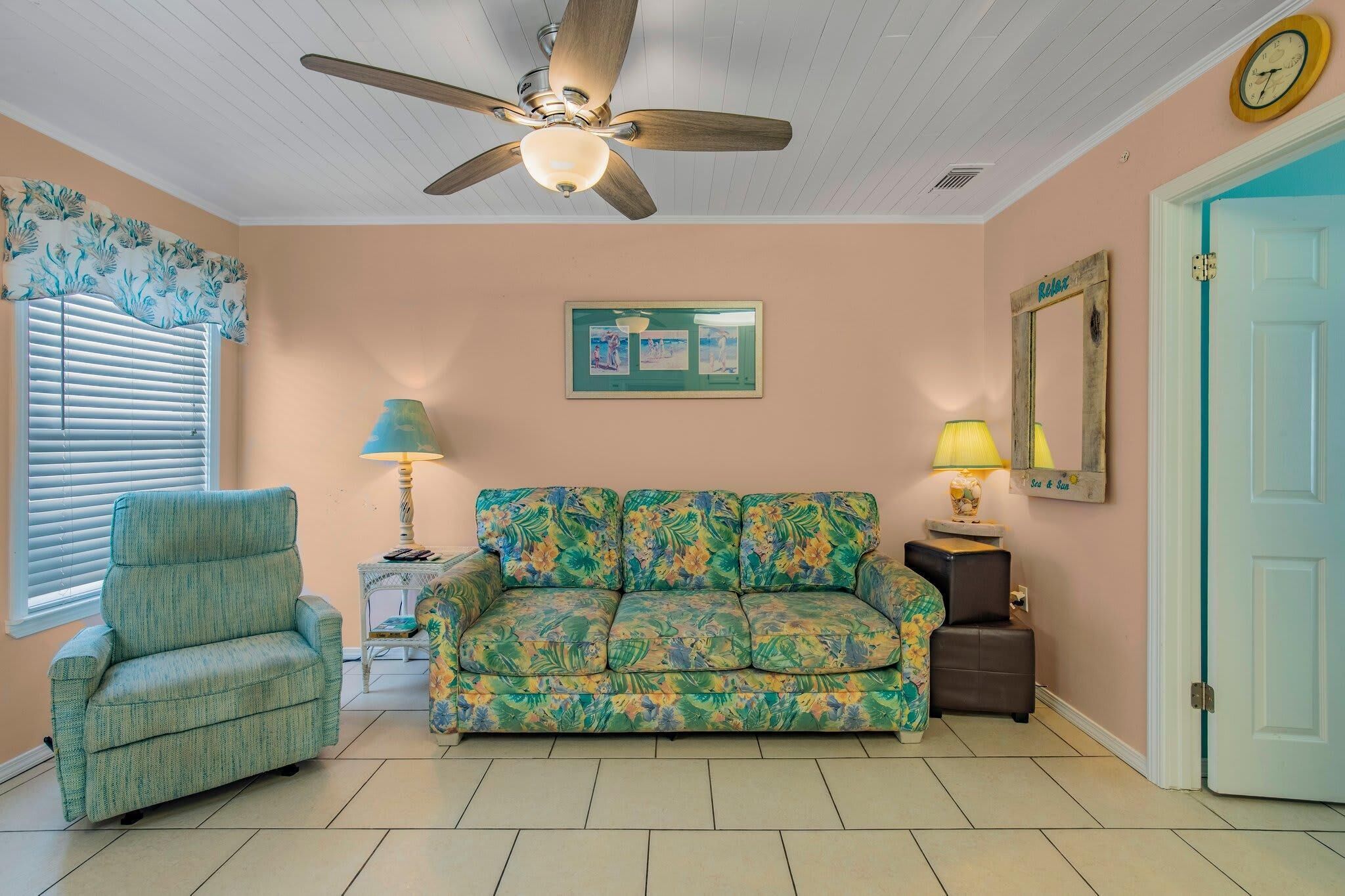 A room with a ceiling fan, a floral sofa, a side chair, a small table with a lamp, a framed picture, and a door with a mirror.