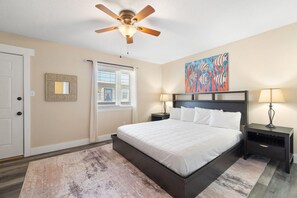 A bedroom with a bed, a ceiling fan, a nightstand with a lamp, a window with blinds, a door, and a painting on the wall. - Gulf View 16 (Gulf Shores)