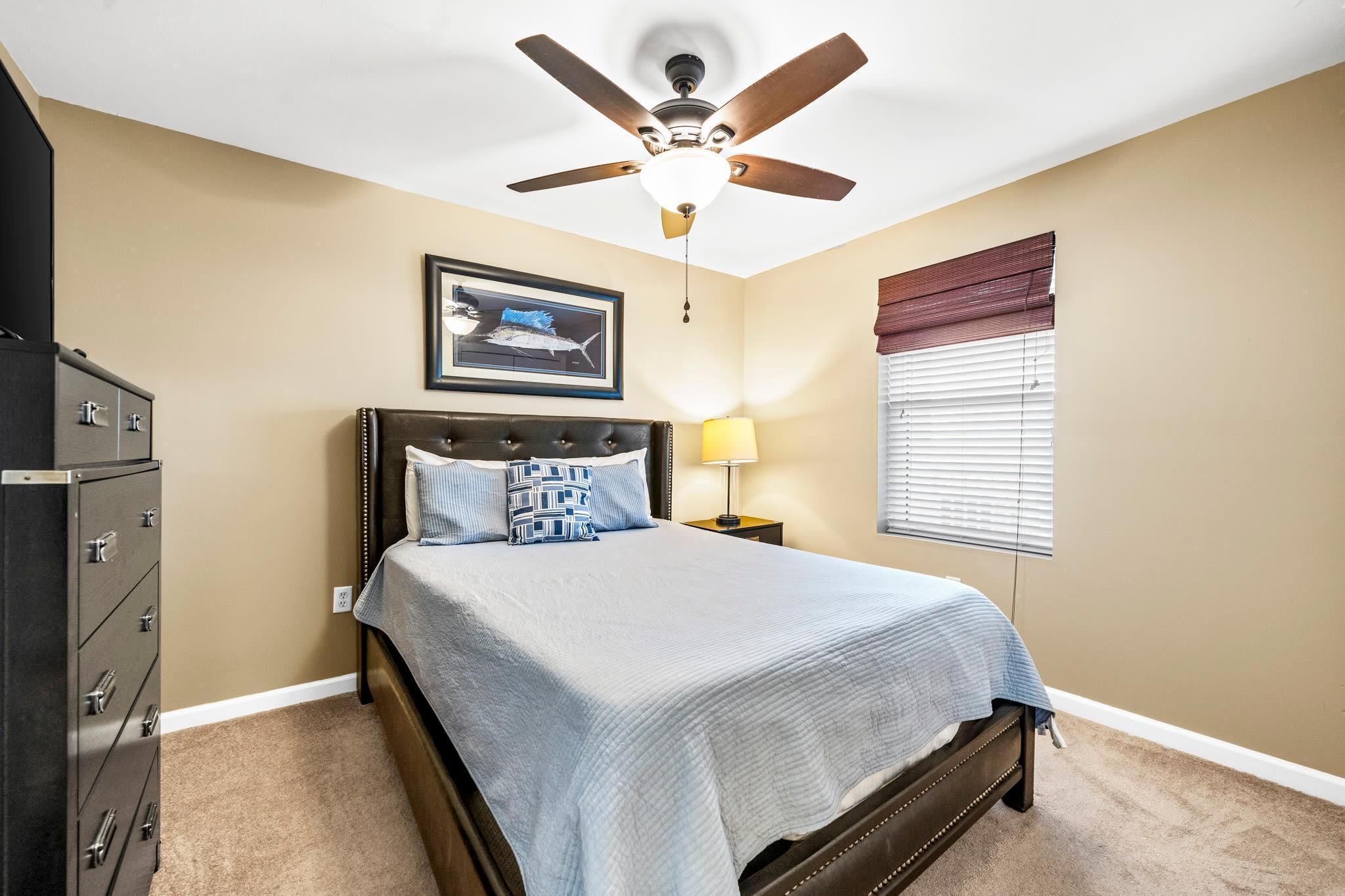A bedroom with a bed, a ceiling fan, a nightstand with a lamp, a dresser, and a framed picture on the wall.