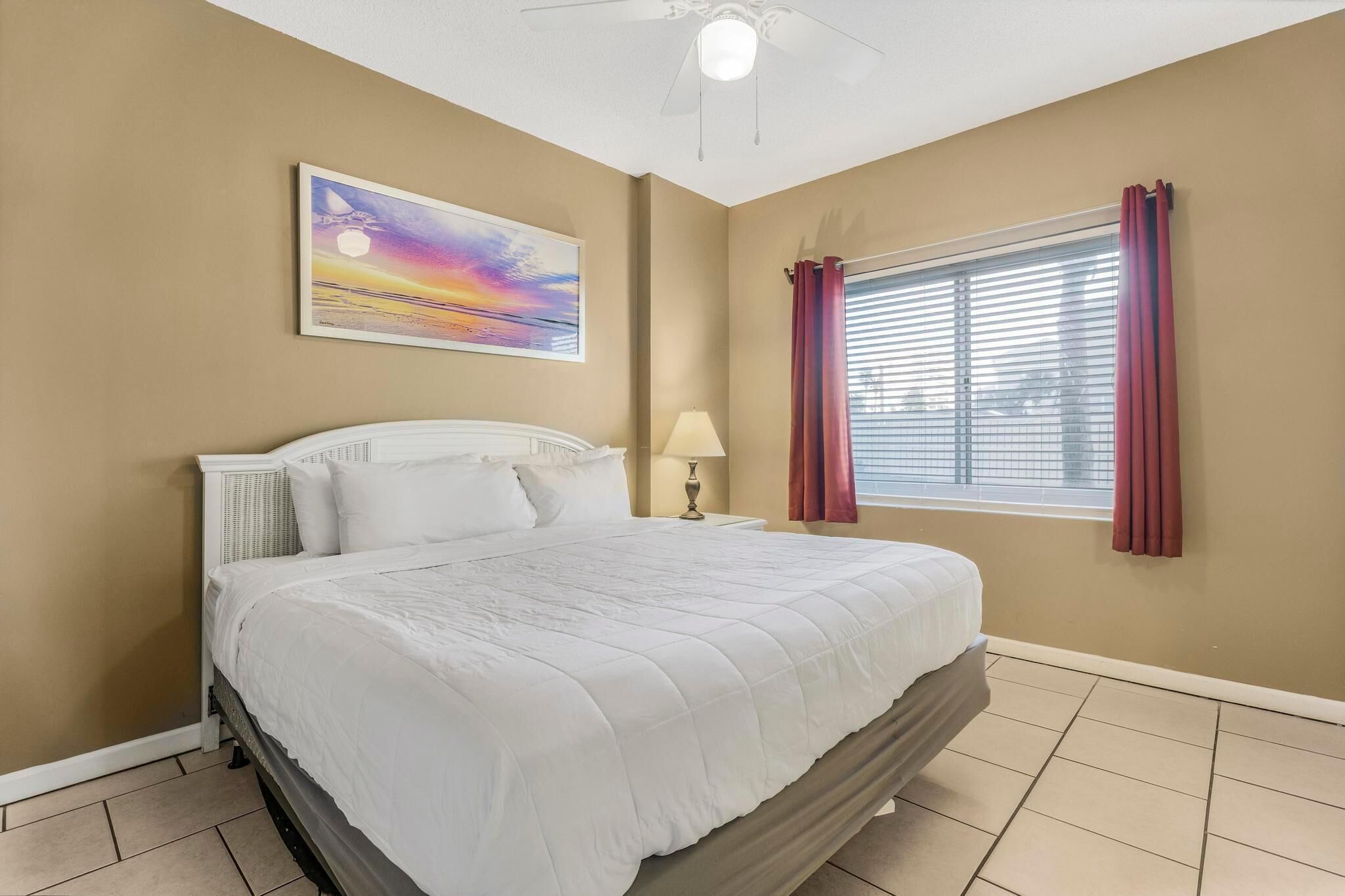 A bedroom with a bed, a nightstand, a lamp, a window with curtains, and a painting on the wall.