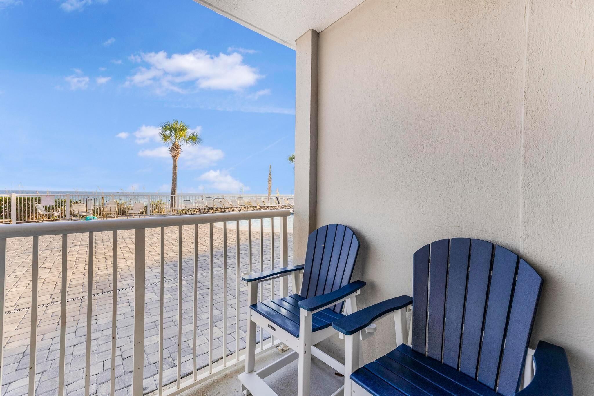 A balcony with two Adirondack chairs, a palm tree, and a view of the beach.