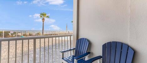 A balcony with two Adirondack chairs, a palm tree, and a view of the beach.