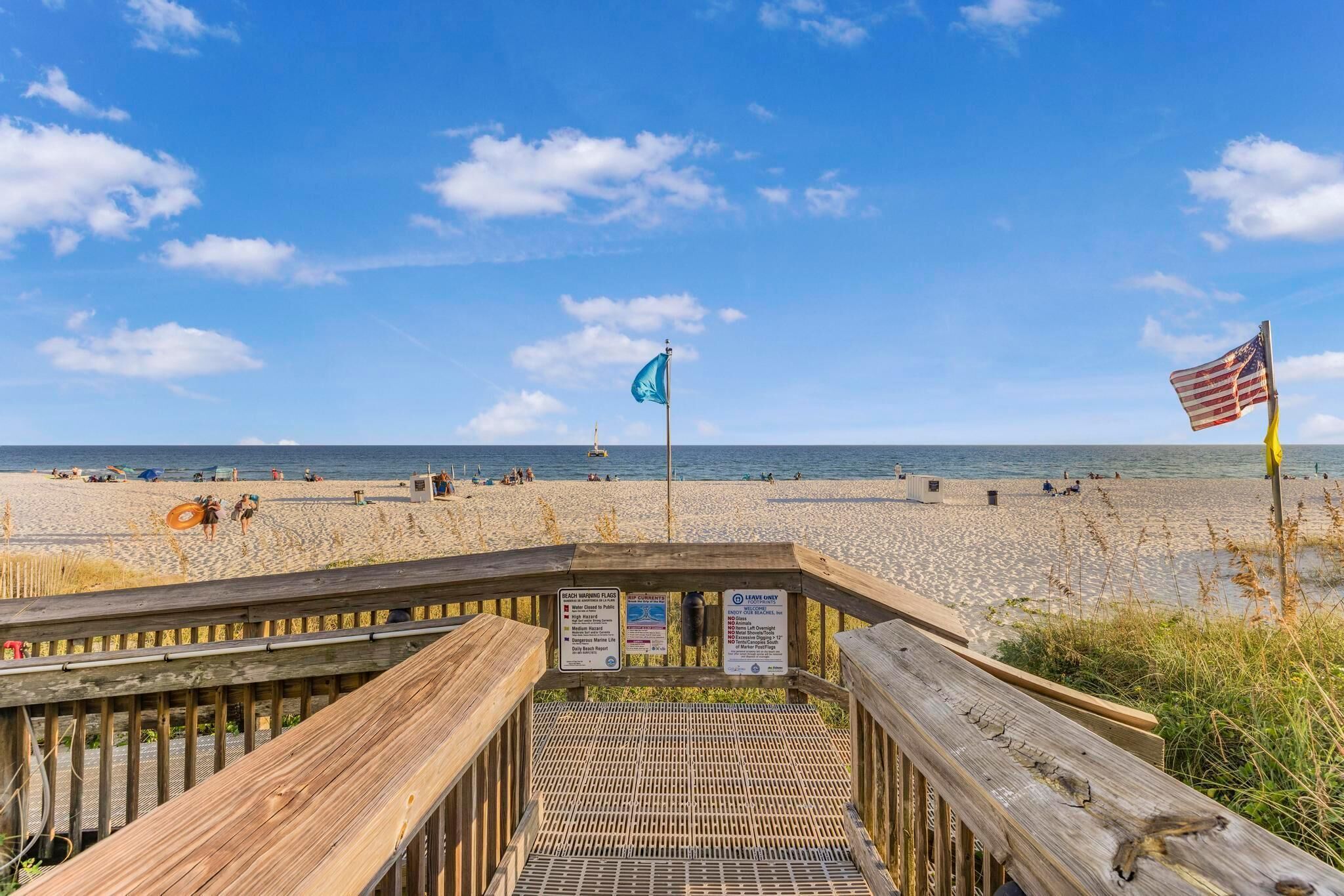 A wooden boardwalk leading to a beach with a blue flag and an American flag.