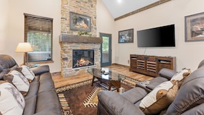 A living room with a stone fireplace, leather sofas, a glass coffee table, and a flat-screen TV mounted on the wall.