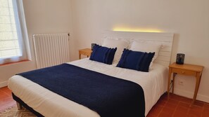 Un lit bien fait, avec une literie blanche et bleue, une table de chevet en bois sur laquelle se trouve une lampe, et un radiateur fixé au mur.