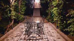 Un chemin pavé mène à une table à plateau de verre, entourée de chaises en métal, le tout étant agrémenté de verdure et avec un escalier en arrière-plan.