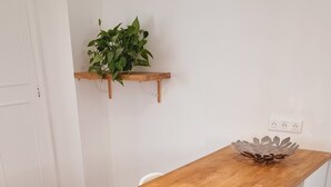 Une table de salle à manger en bois, avec des chaises blanches, une petite plante posée sur une étagère et une prise électrique au mur.