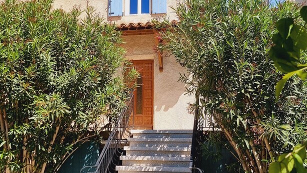 Un bâtiment doté d’un escalier en pierre menant à une porte en bois, entouré de verdure et surplombé d’un ciel bleu clair.