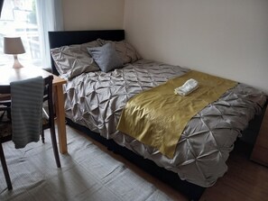A single bed with a grey and gold duvet, a grey pillow, and a gold throw blanket. There is a wooden chair with a grey towel draped over it. A lamp is on a table to the left. - Lovely 1- Double Bed Studio in London (London)