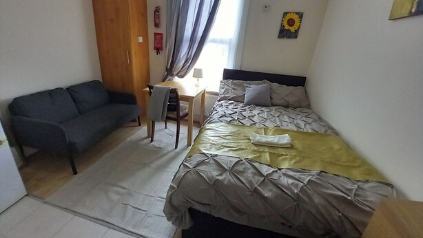 Ein kleiner, gut beleuchteter Raum mit einem Bett, einem Schreibtisch, einem Stuhl und einem Sofa.