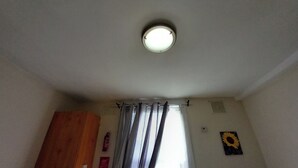 Ein Zimmer mit Deckenleuchte, einem Vorhangfenster, einem Holzschrank und einem Sonnenblumenbild an der Wand.