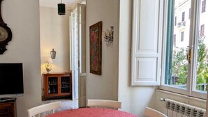 A dining area with a round table covered in a red tablecloth, surrounded by white chairs. There is a window with white shutters, a radiator below, and a television on a stand to the left. - Beautiful 2-bed Apartment in Roma (Rome)