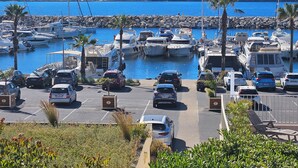 Een jachthaven met aangemeerde boten, een parkeerplaats met auto's en een uitzicht op de zee en heuvels op de achtergrond.