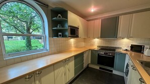 A compact kitchen with white cabinets, a built-in microwave, a stove, and a window overlooking a green garden. - Luxury Apartment In London - Pool Spa & Gym (London)
