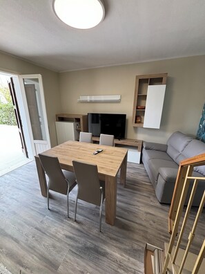 Un salon moderne avec une table à manger en bois, deux chaises, un canapé gris, un téléviseur et des étagères intégrées.
