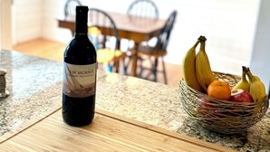 A kitchen with a wine bottle on the counter, a fruit basket, and a dining area in the background. - Ocean view, private 4 Bedroom home just steps to the beach! (Old Orchard Beach)