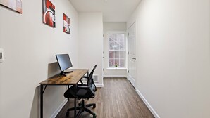 Un pasillo estrecho con un escritorio de madera, una silla de oficina negra y una computadora. Hay tres cuadros abstractos en la pared. Se ve una ventana con vista a un edificio al fondo.