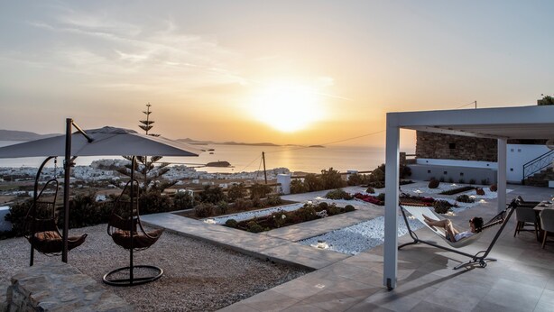 A modern outdoor seating area with a view of the sunset over the sea, including a hammock and two swing chairs. - Modern Cycladic House (Naxos)