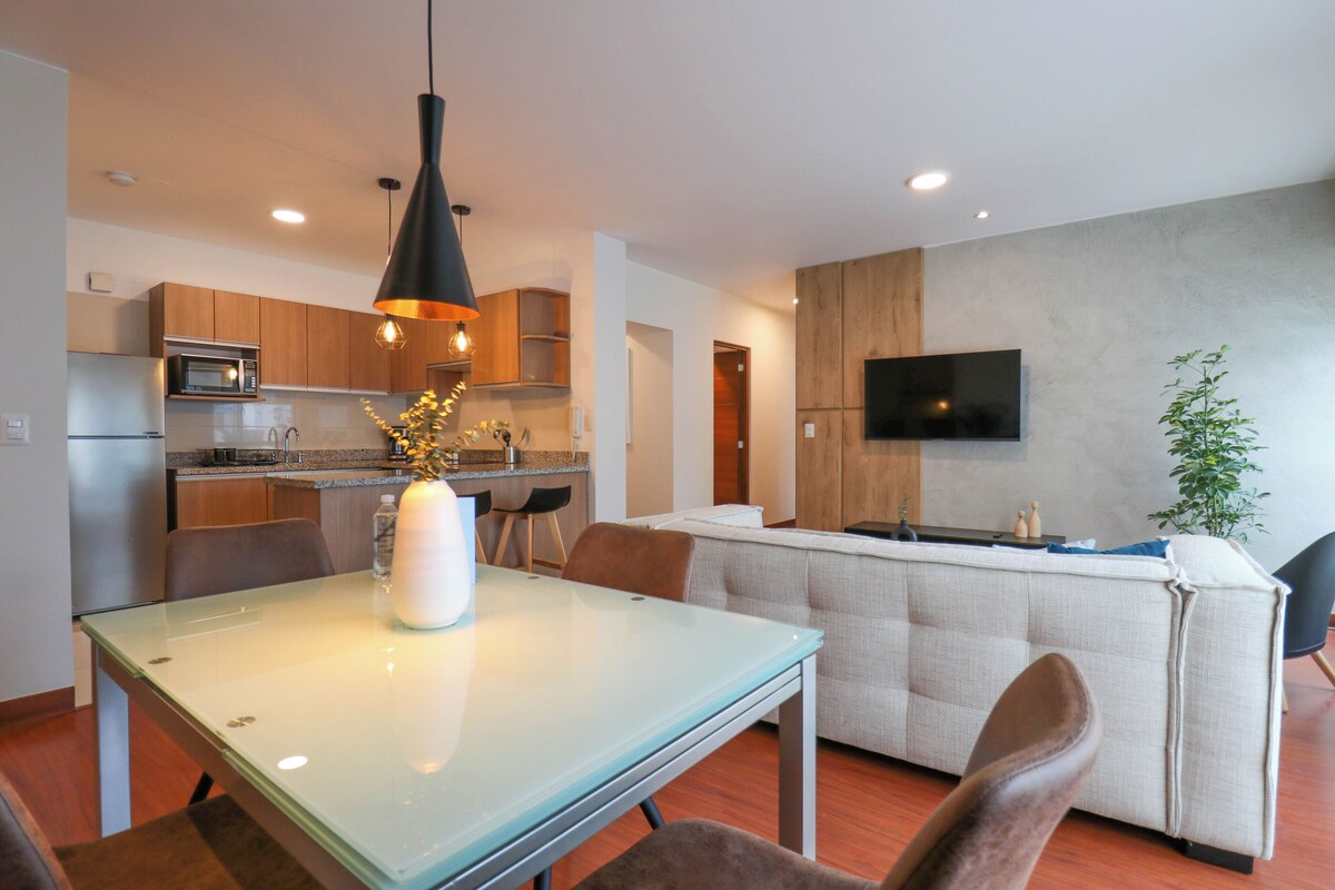 Una cocina moderna con armarios de madera, microondas integrado y un comedor con mesa y sillas de vidrio.