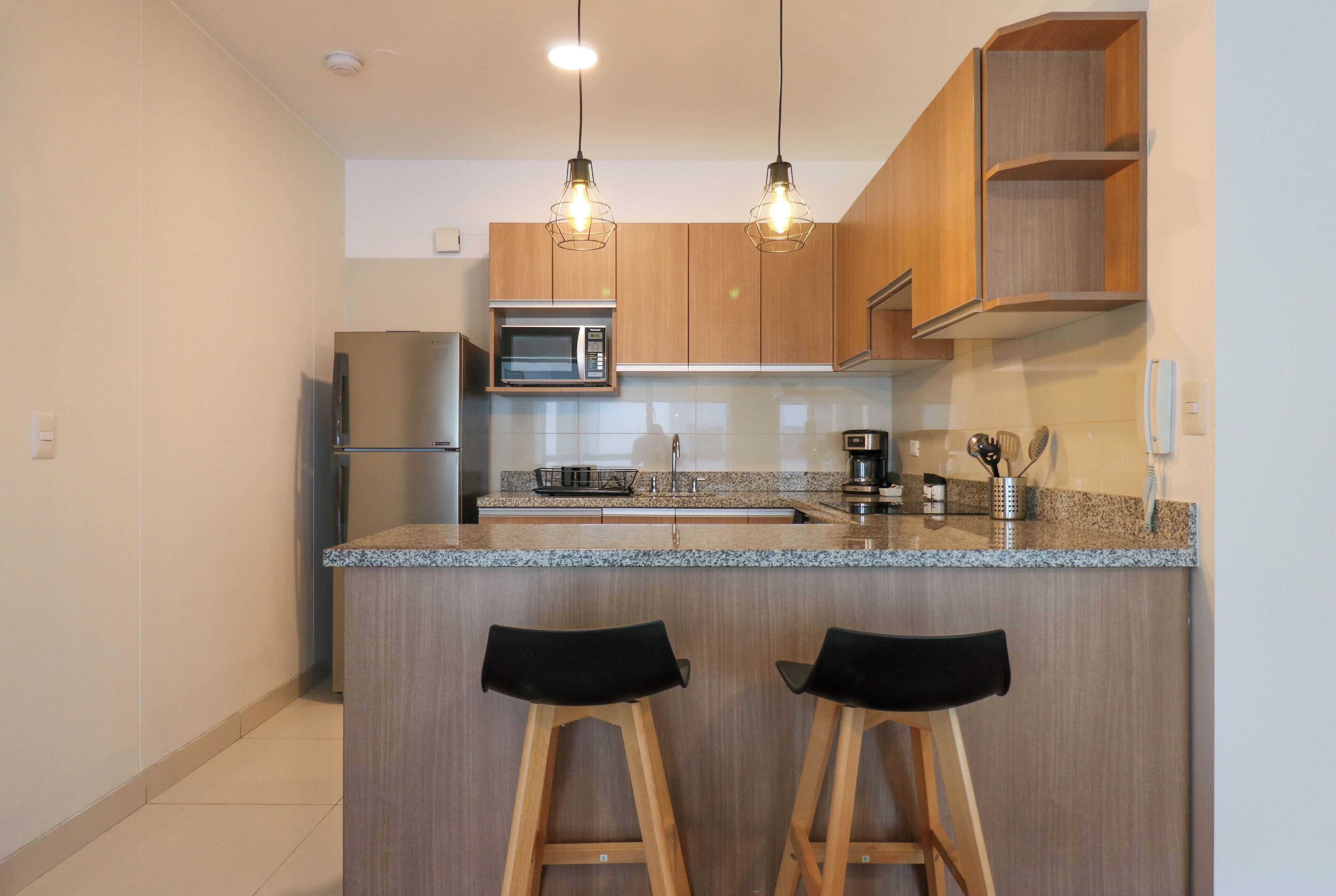 A modern kitchen with wooden cabinets, a granite countertop, two bar stools, and pendant lighting.