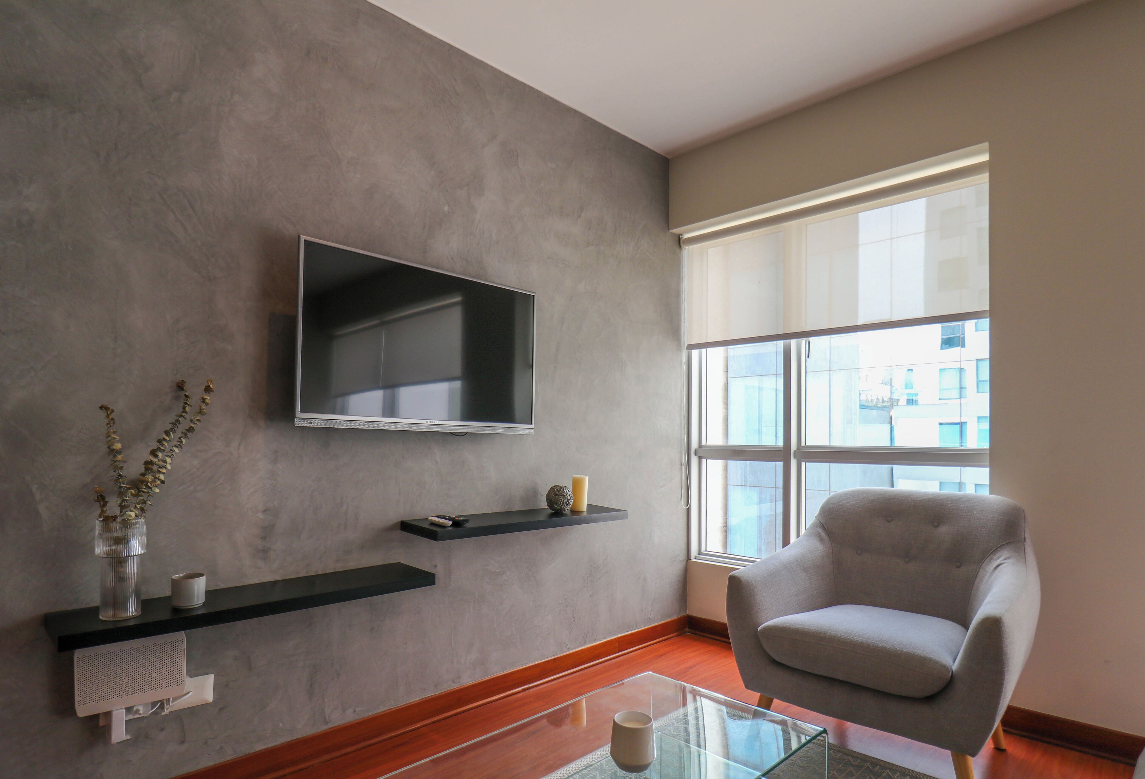 A modern living room with a flat-screen TV mounted on a textured wall, a grey armchair, and a glass coffee table.
