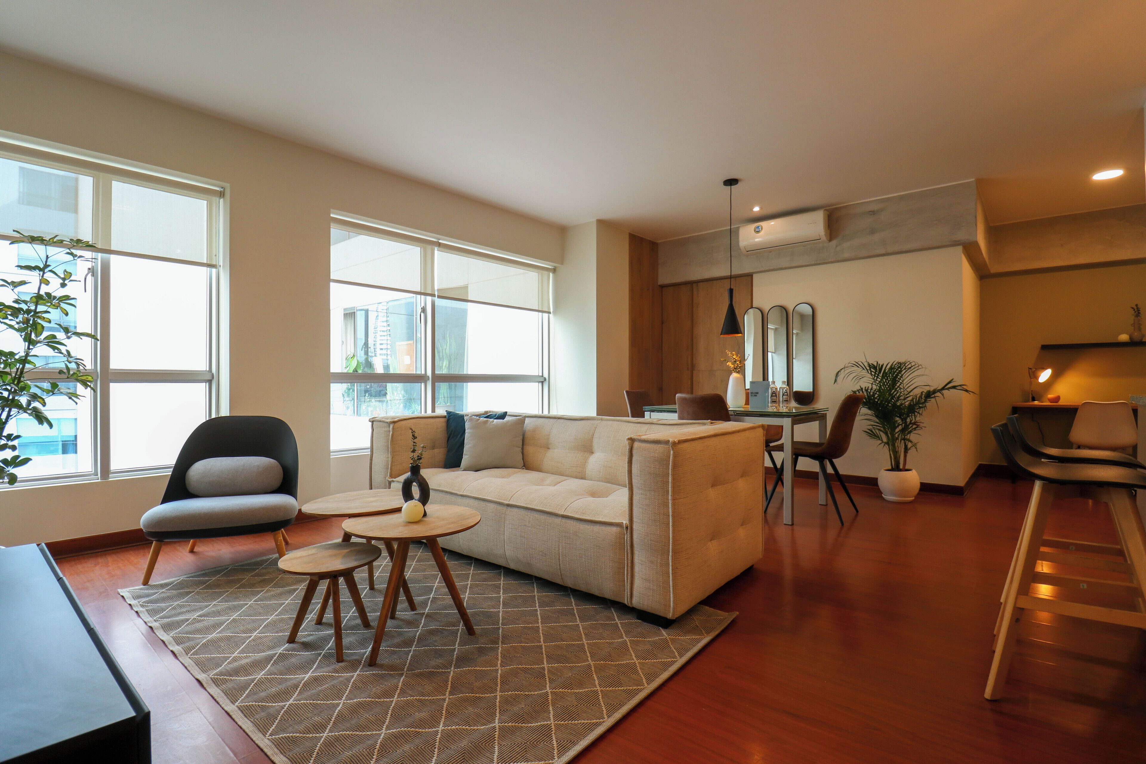 Un salon moderne avec un canapé, une table basse et des fauteuils. On aperçoit à l’arrière-plan un coin cuisine avec une table à manger et des chaises.