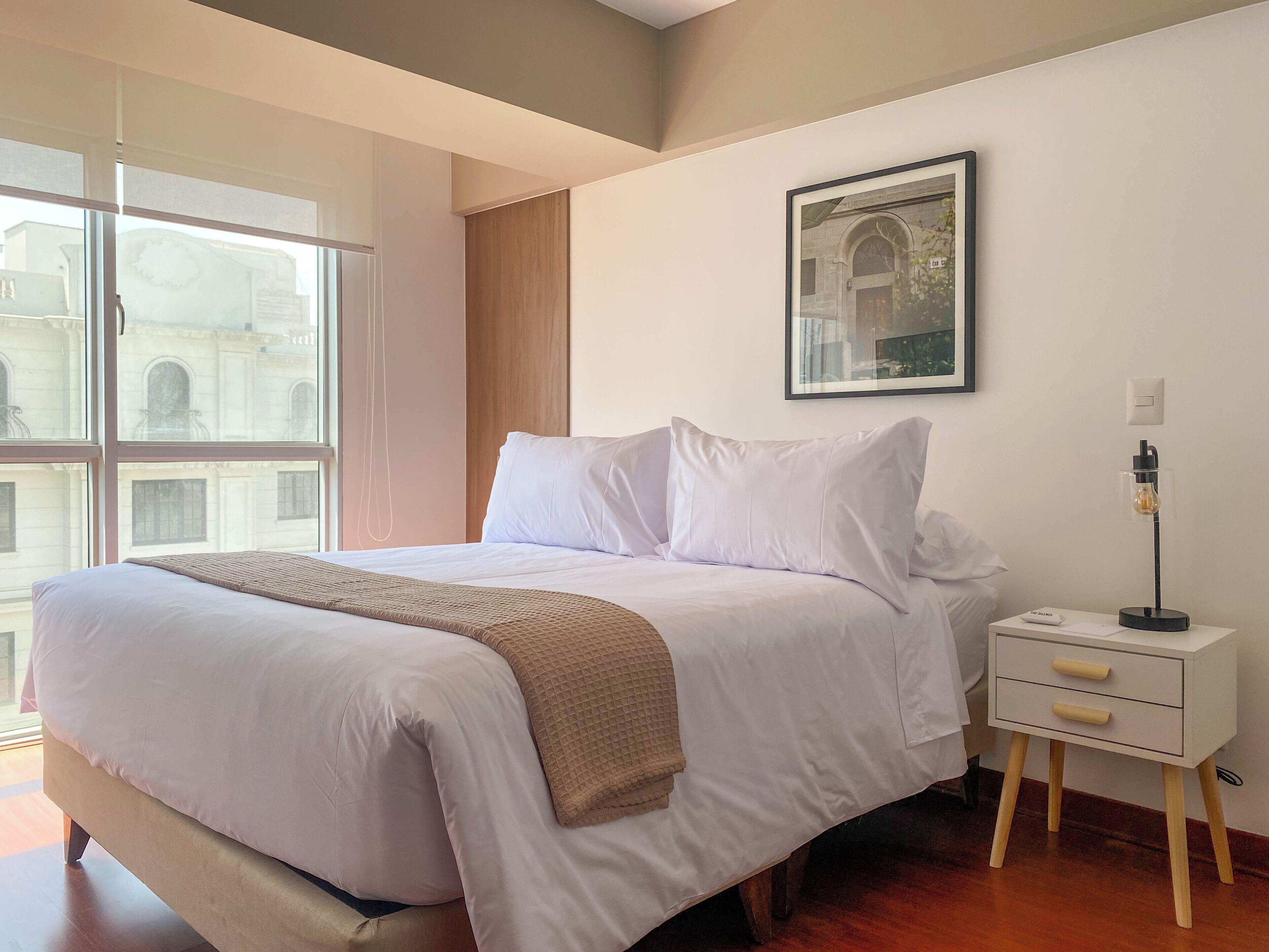 A neatly made bed with white linens and a beige throw blanket, a bedside table with a lamp, and a framed picture on the wall.