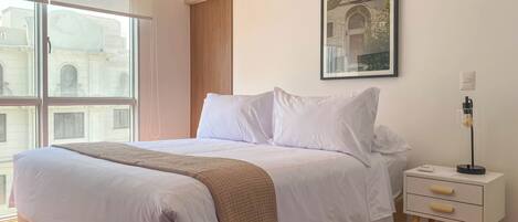 A neatly made bed with white linens and a beige throw blanket, a bedside table with a lamp, and a framed picture on the wall.