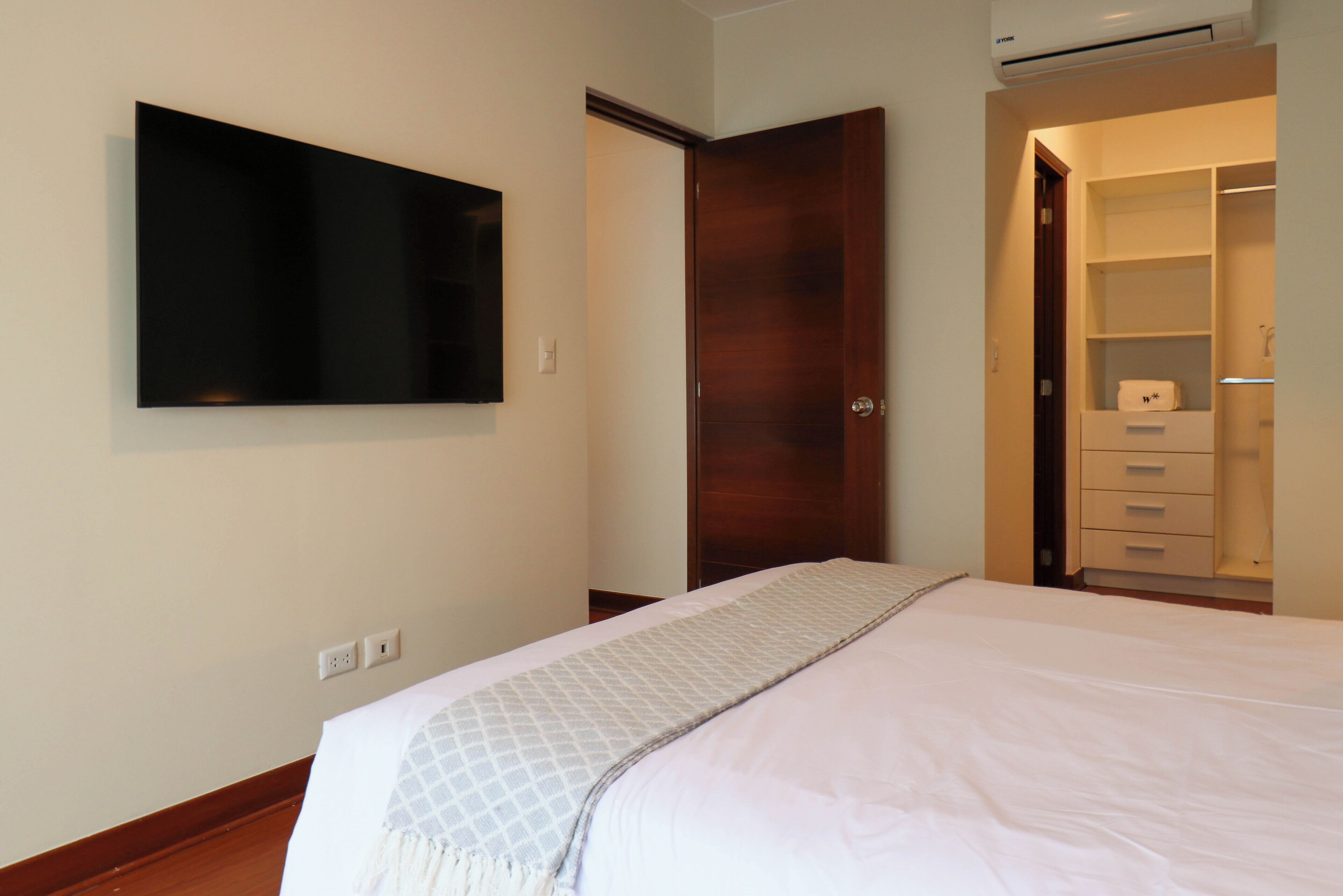A bedroom with a bed, a wardrobe, a television mounted on the wall, and a door leading to a bathroom.