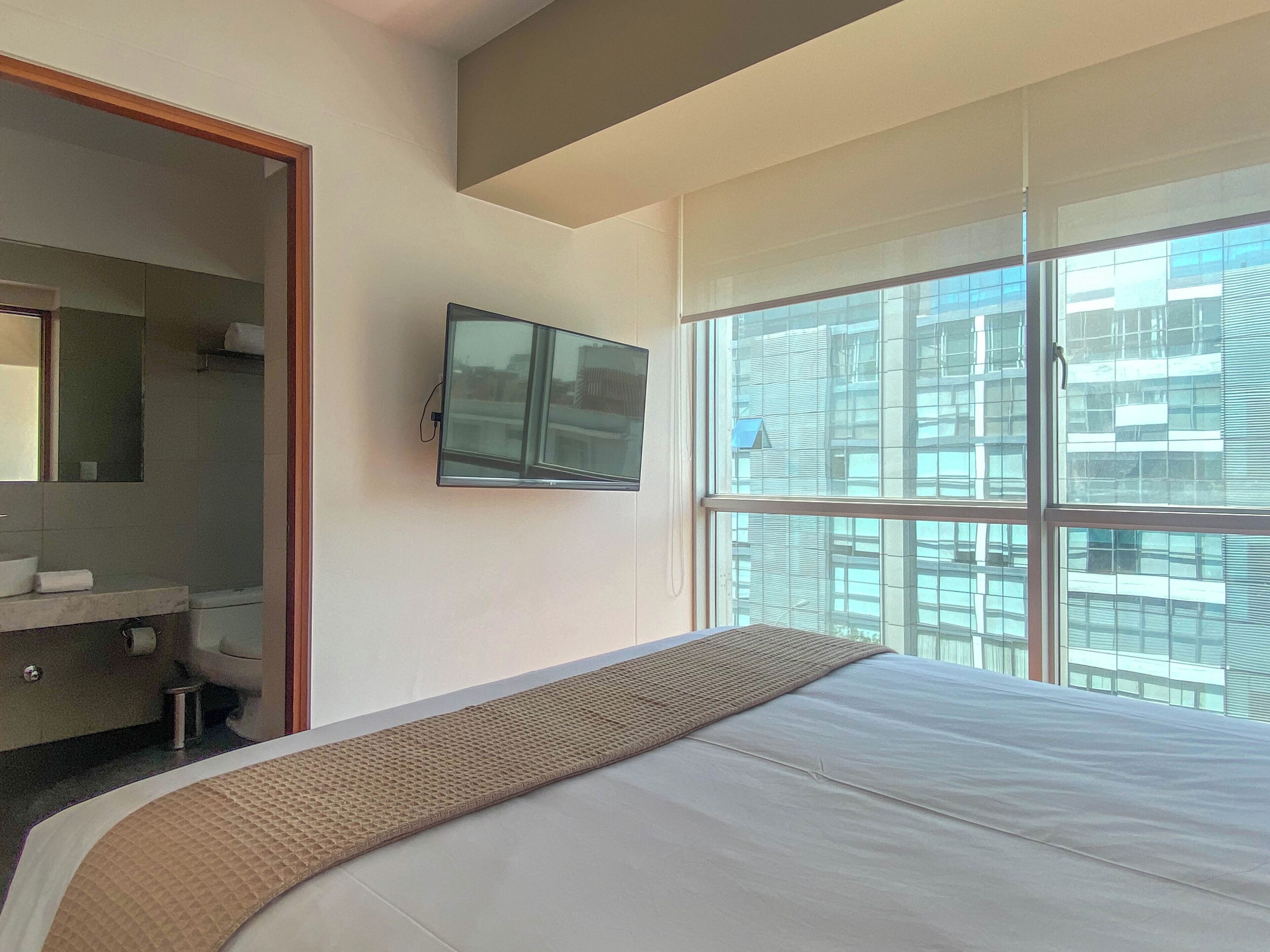 A modern hotel room with a large bed, a television mounted on the wall, a bathroom with a mirror and sink, and a view of the city through large windows.